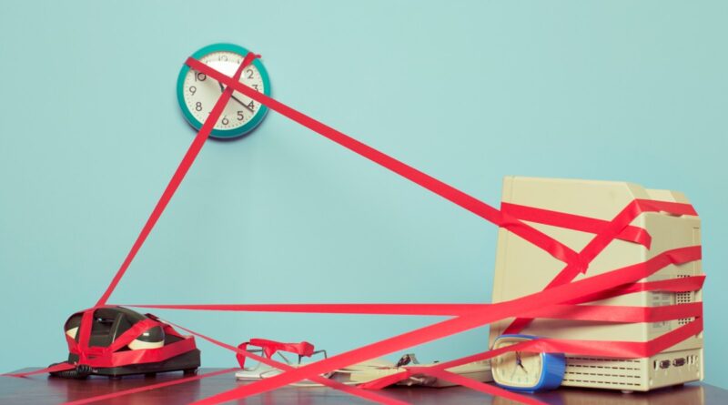 Image of a computer, phone and clock on a desk tied in red tape.